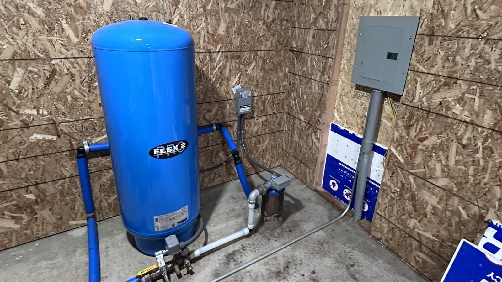 Subpanel mounted in a detached pump house with conduit running to it and a blue well pressure tank beside it