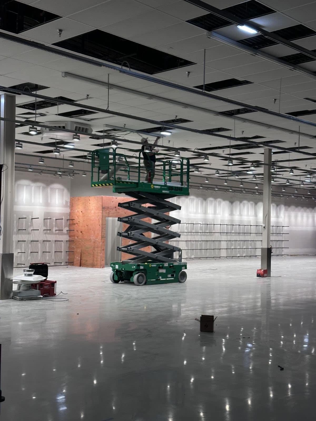 Electrician on a scissor lift installing the sales-floor lighting package into the suspended ceiling grid of a retail tenant improvement