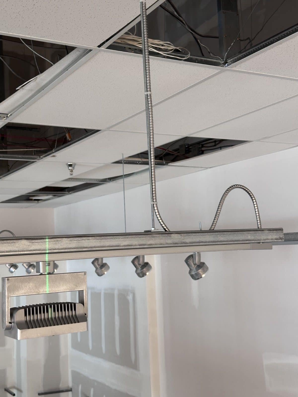 Close-up of a retail track lighting run fed by an MC whip and supported on threaded rod, aligned to a green laser layout line
